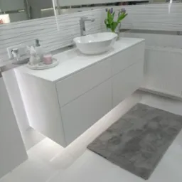 Nowoczesna, biała szafka łazienkowa pod umywalkę nablatową, z subtelnym oświetleniem LED od dołu, uzupełniona o dekoracje: ceramicznego zajączka, świecę i wazon z fioletowymi tulipanami.