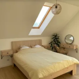 Jasna sypialnia na poddaszu z drewnianym łóżkiem, szafkami nocnymi na czarnych wspornikach i okrągłym lustrem nad toaletką, oświetlona lampą z abażurem z wikliny i oknem dachowym.