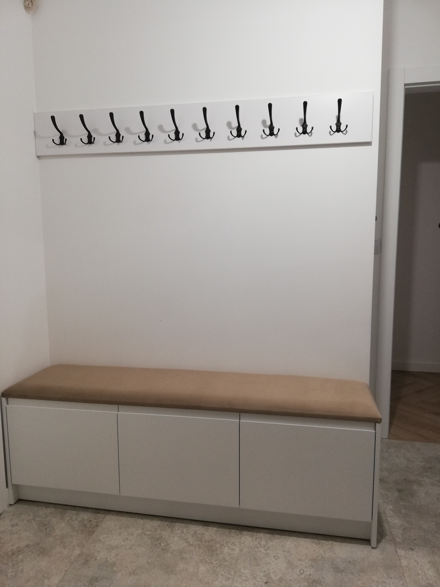 Wnętrze przedpokoju z białą szafką z tapicerowanym siedziskiem i wieszakiem z czarnymi haczykami na białej listwie.
