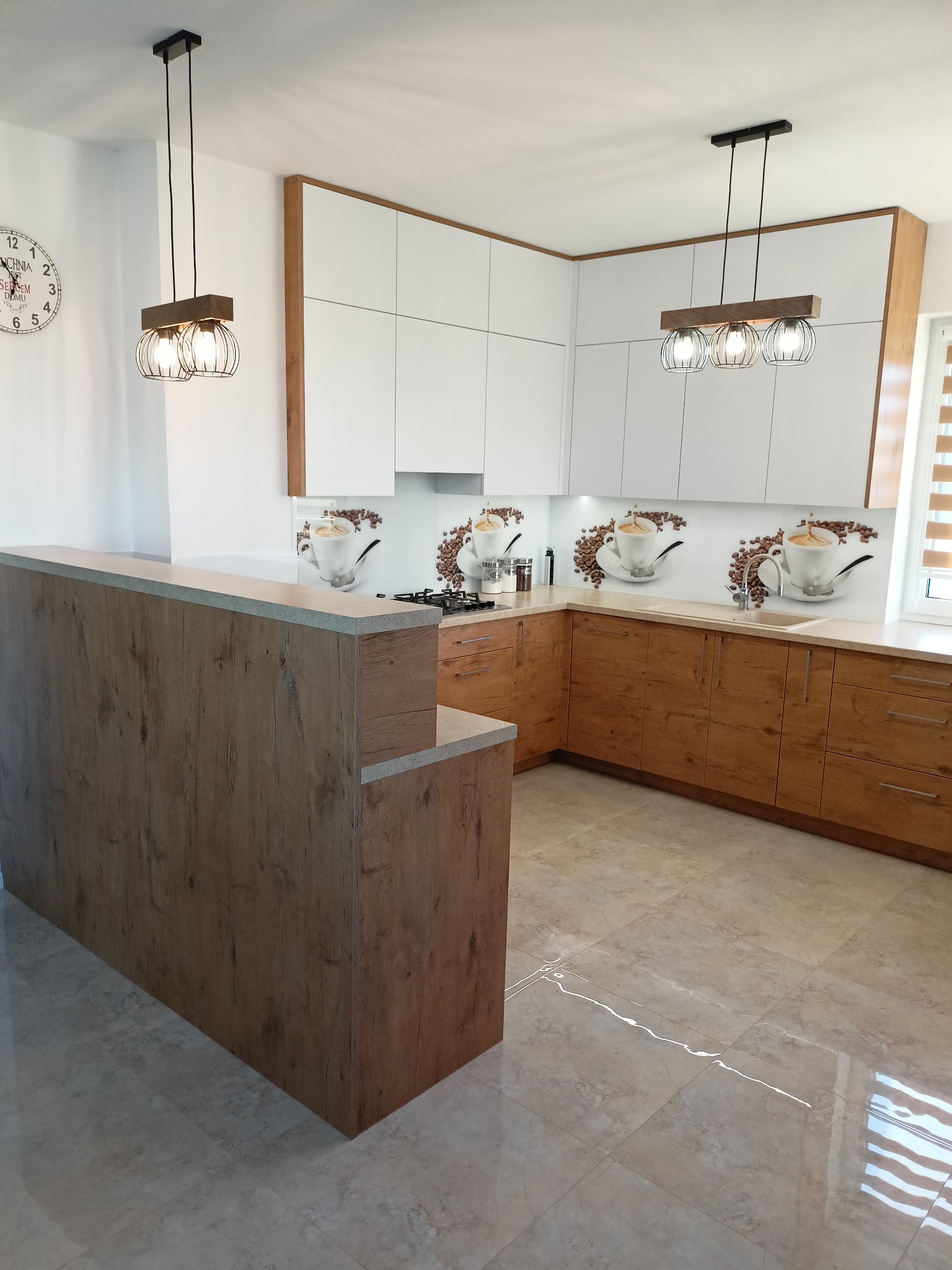 Nowoczesna kuchnia z drewnianymi i białymi szafkami, blatem w kolorze jasnego kamienia i dekoracyjnym panelem szklanym z motywem kawy, oświetlona lampami wiszącymi z metalowymi kloszami.