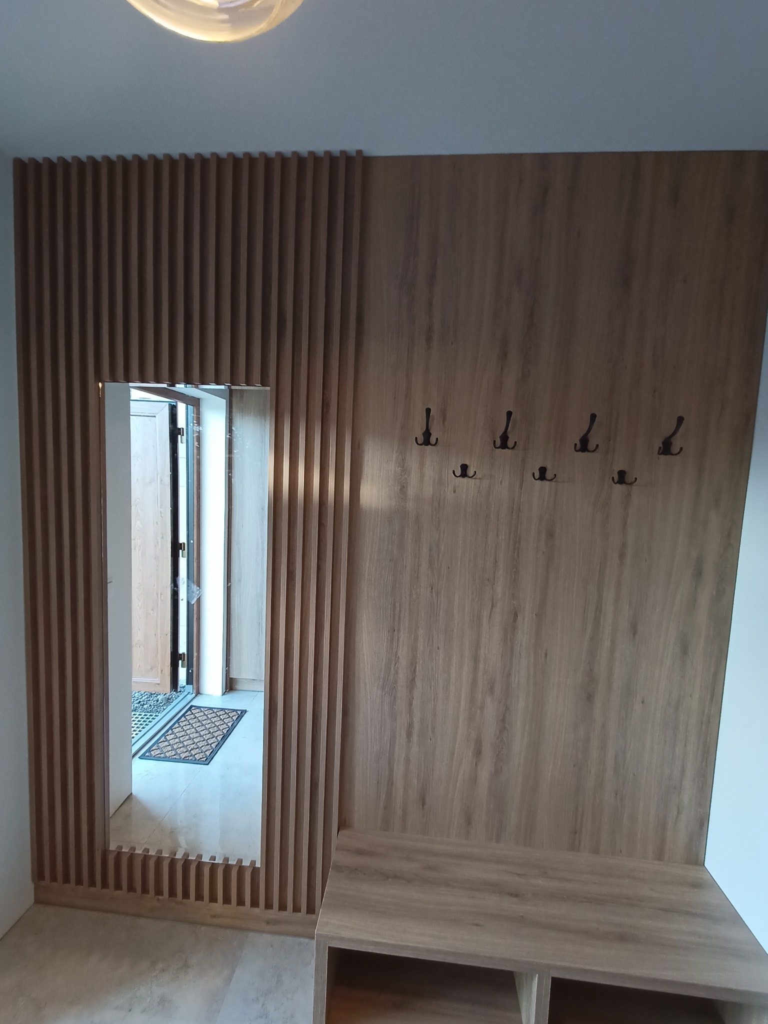 Drewniana zabudowa przedpokoju z lustrem w formie pionowych listewek, w tle widoczna wycieraczka przed drzwiami wejściowymi.