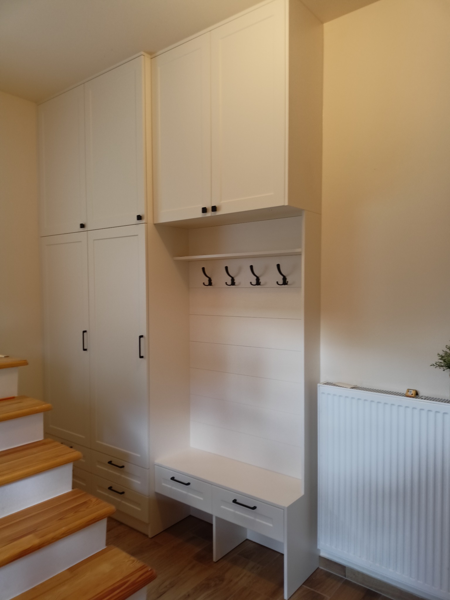 Biała zabudowa przedpokoju z szafami, siedziskiem i wieszakami, umieszczona przy drewnianych schodach