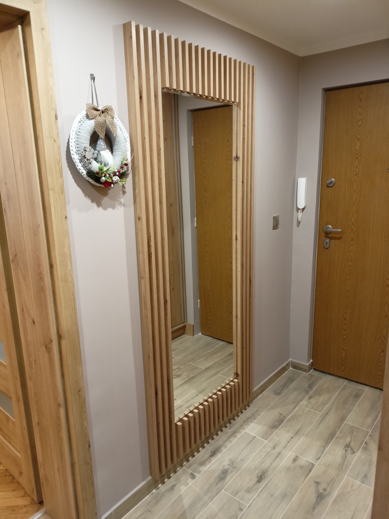 Nowoczesne lustro w drewnianej ramie z pionowych listewek, wiszący wianek świąteczny na ścianie, drewniane drzwi w tle, płytki podłogowe imitujące drewno.