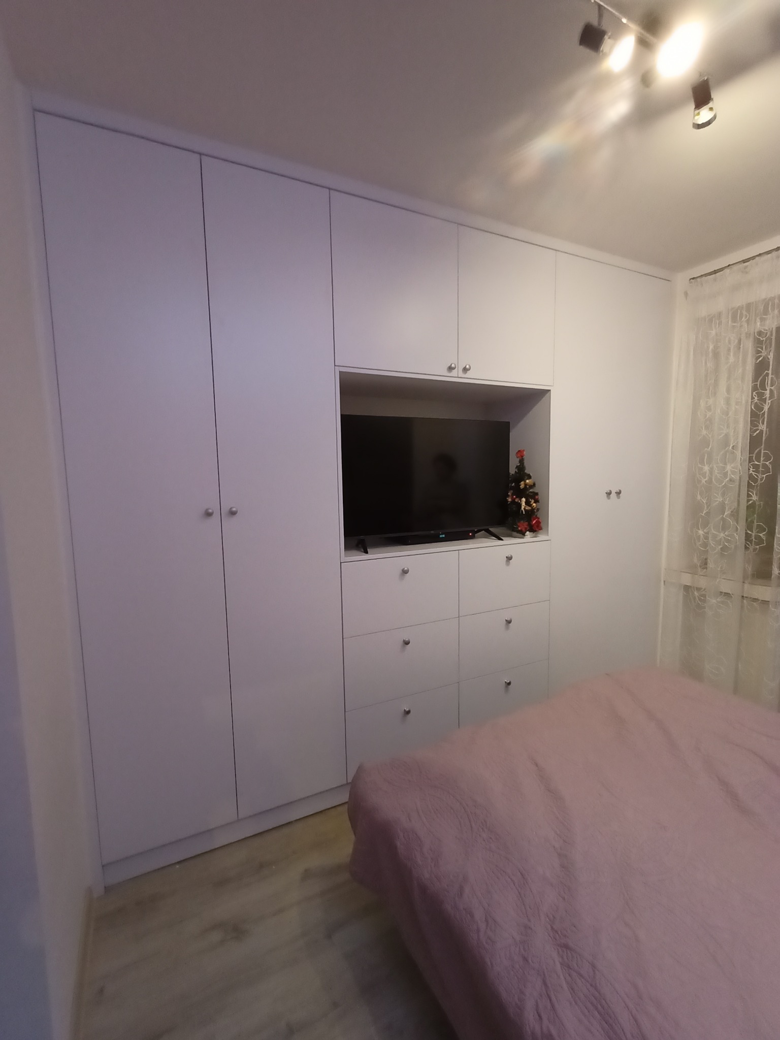 Biała szafa wnękowa z wbudowanym miejscem na telewizor i komodą z szufladami, w sypialni z łóżkiem i choinką dekoracyjną na półce.