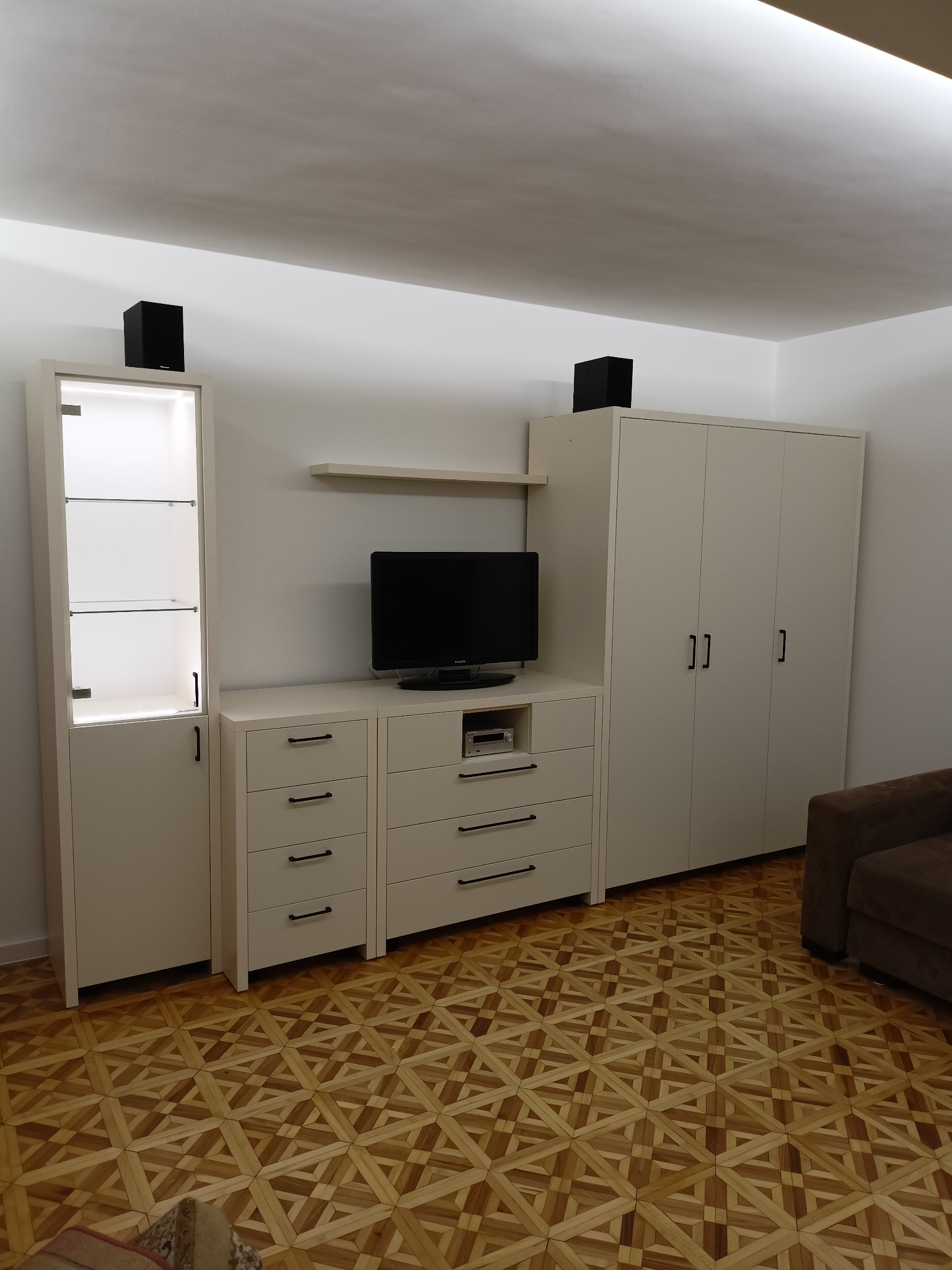 Biała zabudowa meblowa w salonie z szafą, komodą, witryną i półką, z czarnymi głośnikami na górze i parkietem w jodełkę na podłodze.