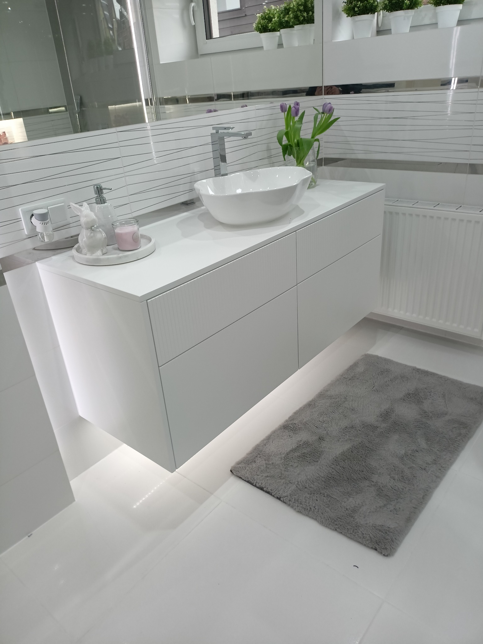 Nowoczesna, biała szafka łazienkowa pod umywalkę nablatową, z subtelnym oświetleniem LED od dołu, uzupełniona o dekoracje: ceramicznego zajączka, świecę i wazon z fioletowymi tulipanami.