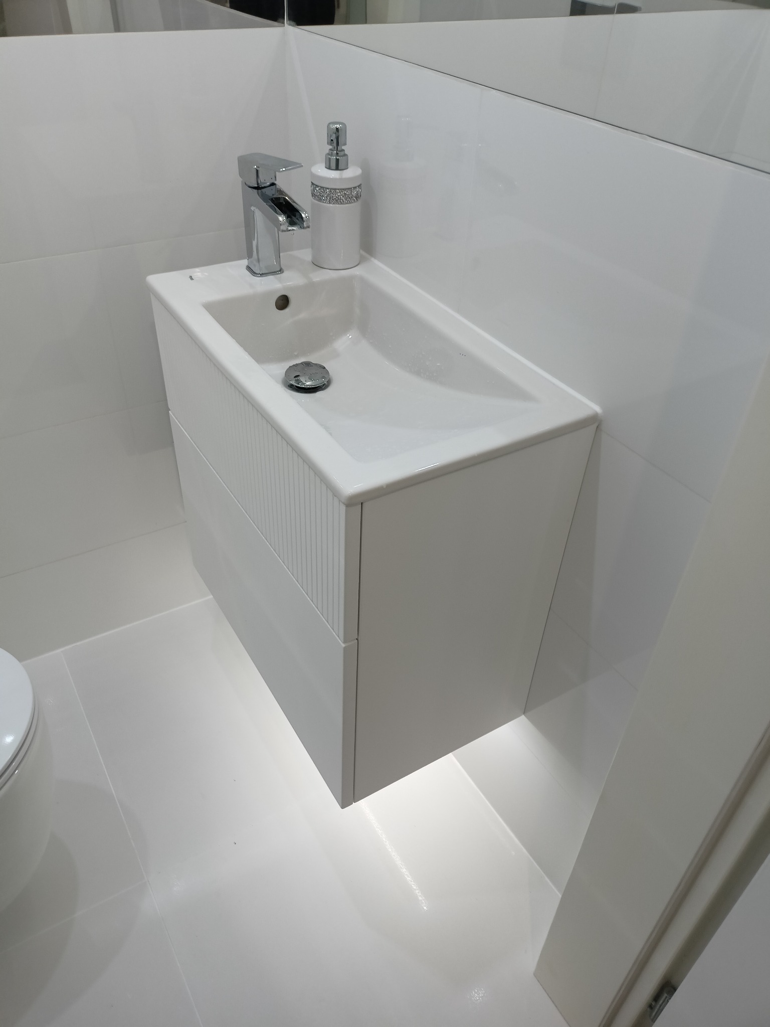 Nowoczesna, biała umywalka z szafką podwieszaną, chromowana bateria, dekoracyjny dozownik mydła, oświetlenie LED pod szafką, minimalistyczna łazienka.