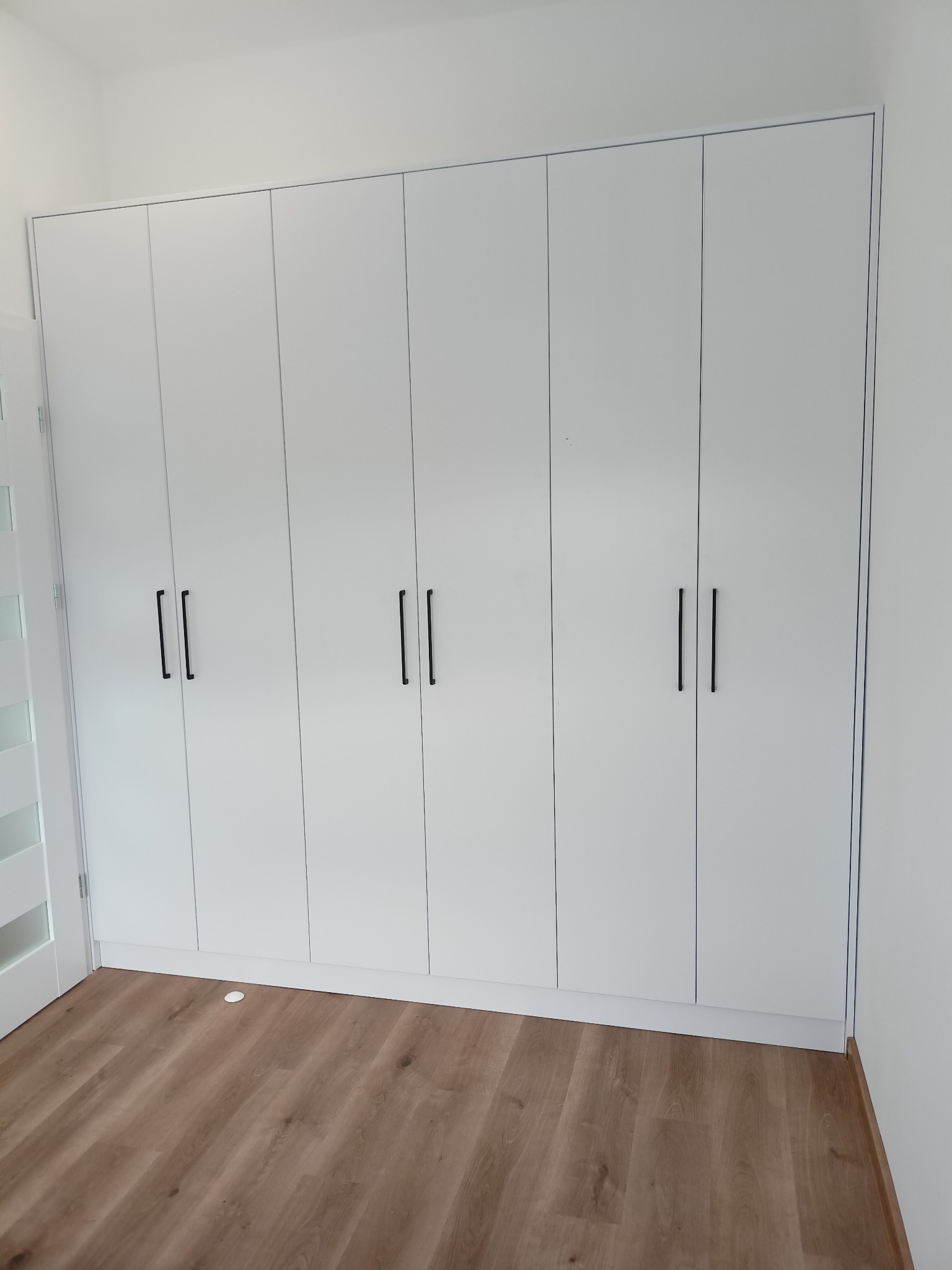 Biała szafa wnękowa z sześcioma drzwiami i długimi, czarnymi uchwytami, na podłodze panele imitujące drewno, widoczny fragment białej ściany i drzwi z szybą.