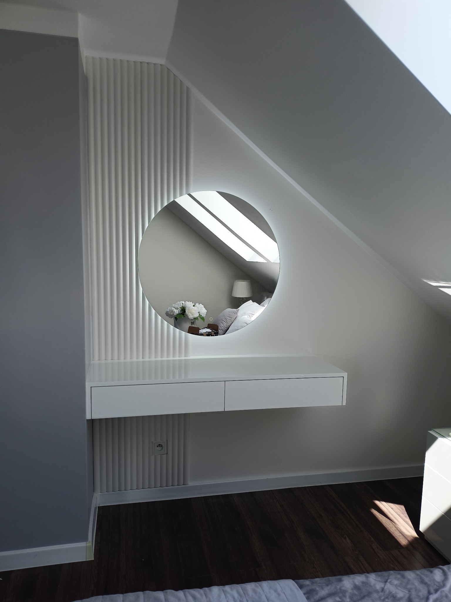 Biała toaletka z okrągłym lustrem podświetlanym LED w pokoju na poddaszu, widoczne dekoracyjne panele ścienne i odbicie sypialni w lustrze.