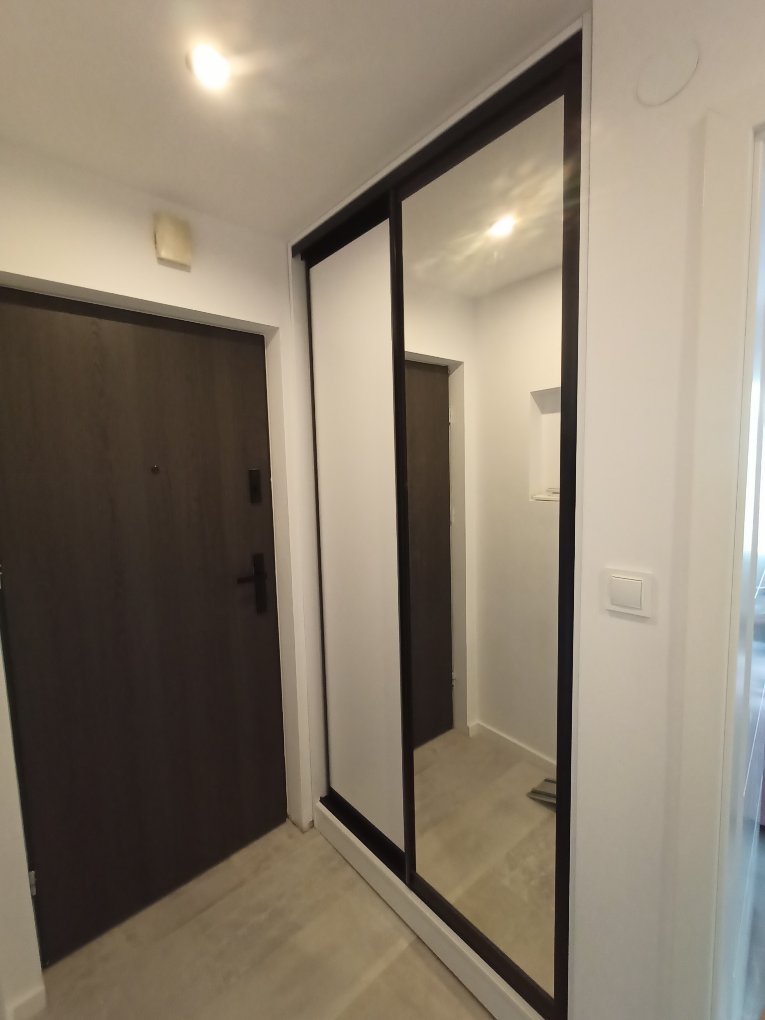 Nowoczesna szafa wnękowa z przesuwnymi drzwiami lustrzanymi i białymi panelami bocznymi, widoczna w korytarzu obok ciemnych drzwi wejściowych.