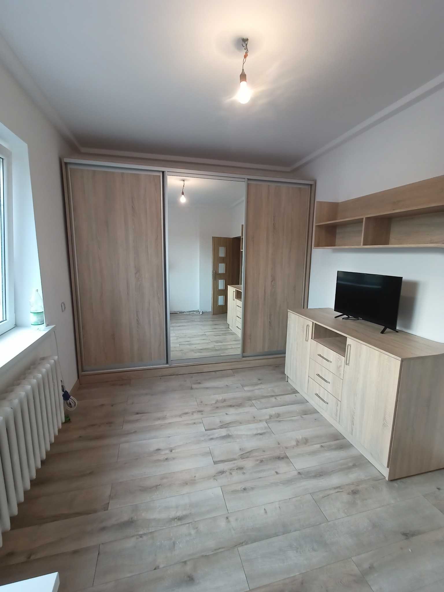 Jasna szafa wnękowa z przesuwnymi drzwiami lustrzanymi, komoda z telewizorem i półki na ścianie w pokoju z panelami podłogowymi imitującymi drewno.