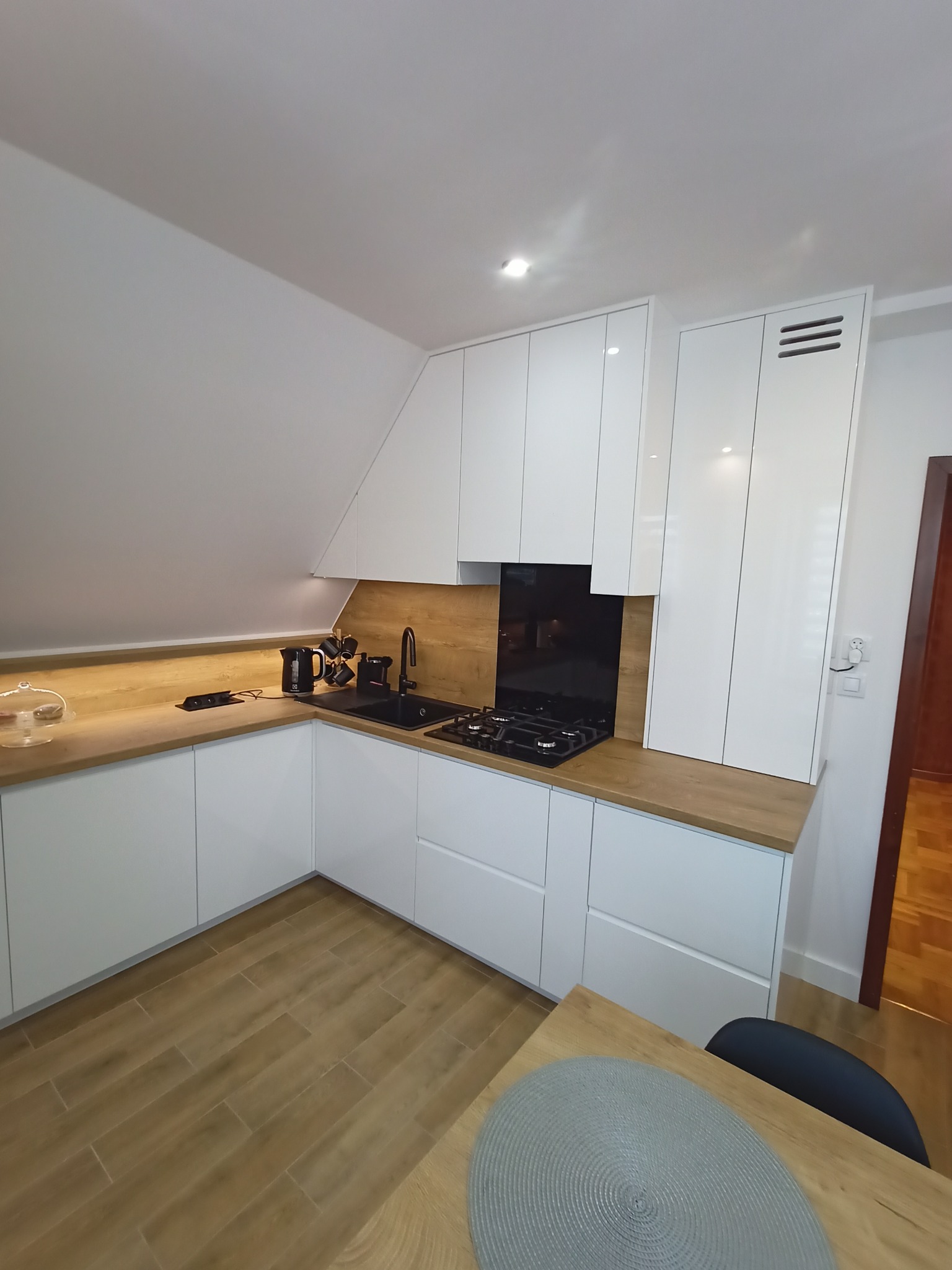 Nowoczesna kuchnia w skandynawskim stylu z białymi szafkami, drewnianym blatem i czarną armaturą, oświetlona światłem punktowym pod skośnym sufitem.