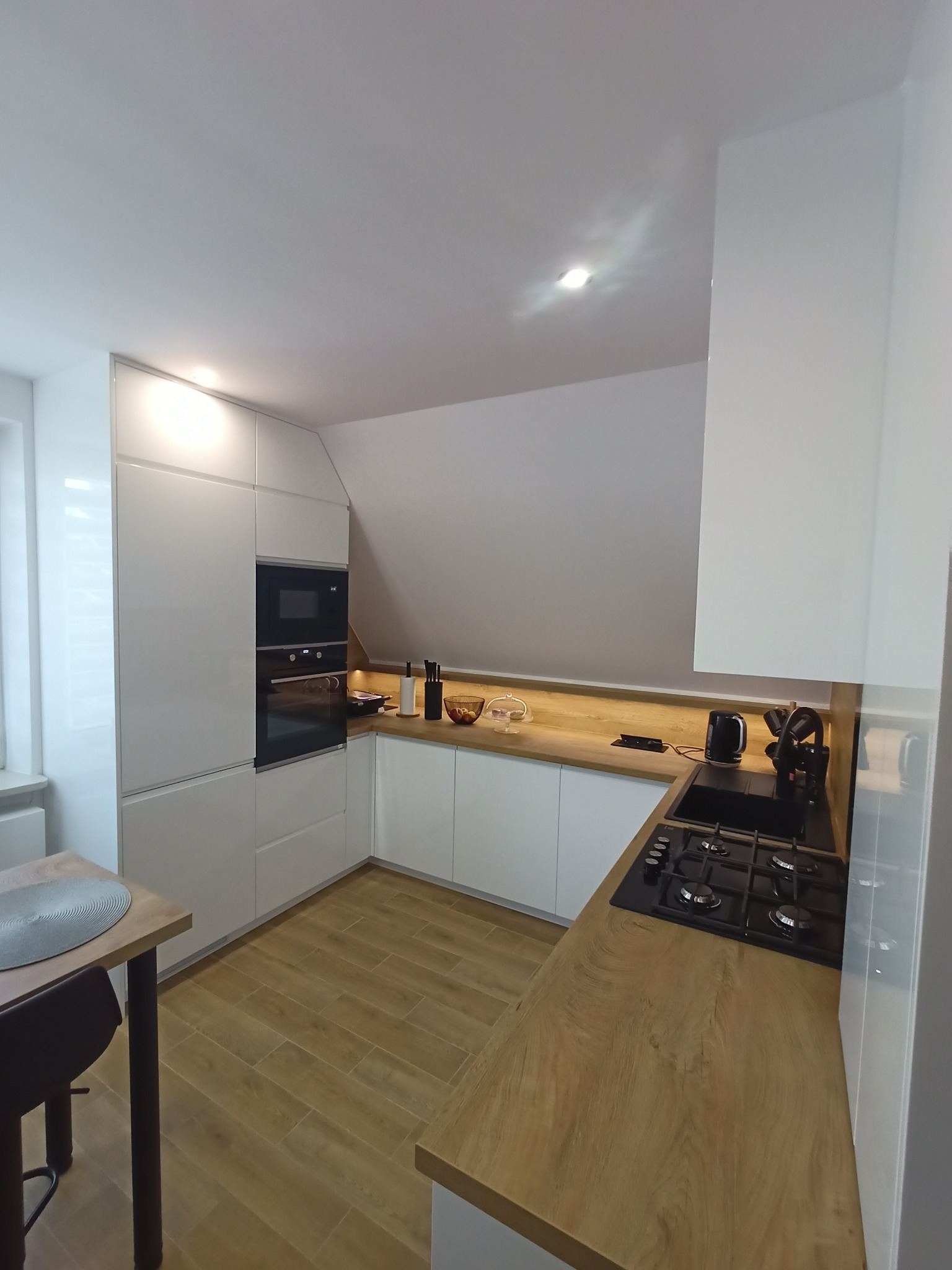 Nowoczesna kuchnia z białymi szafkami, drewnianym blatem i płytą gazową w narożnym układzie, oświetlenie LED podszafkowe, na podłodze panele imitujące drewno, zlew wpuszczony w blat.