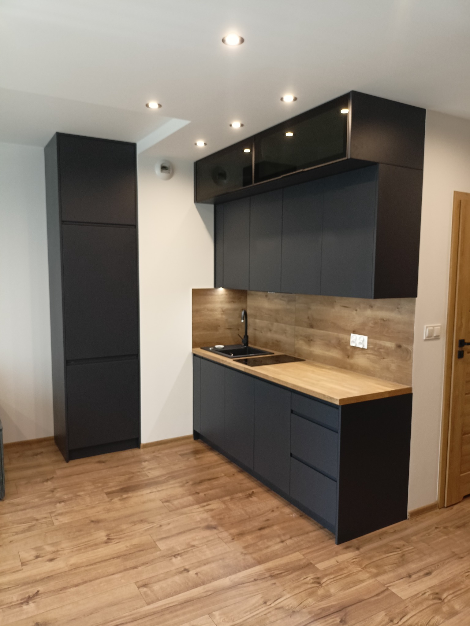 Nowoczesna, minimalistyczna kuchnia w odcieniach szarości z drewnianym blatem i pasującą okładziną ścienną, oświetlona punktowymi lampami sufitowymi i uzupełniona drewnianą podłogą.