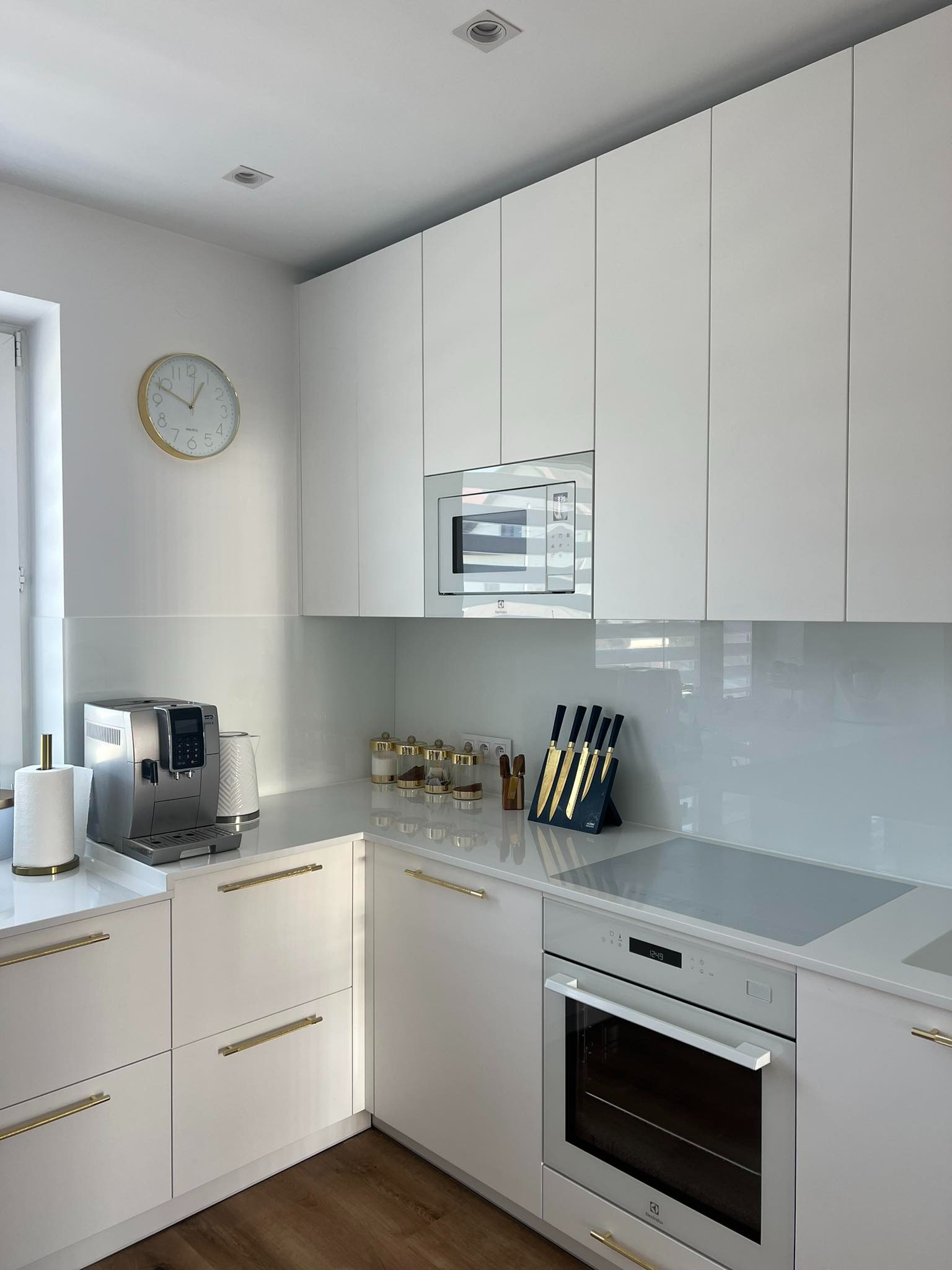 Minimalistyczna, jasna kuchnia z białymi, lakierowanymi frontami, złotymi uchwytami, sprzętem AGD marki Electrolux i złotym zegarem na ścianie.