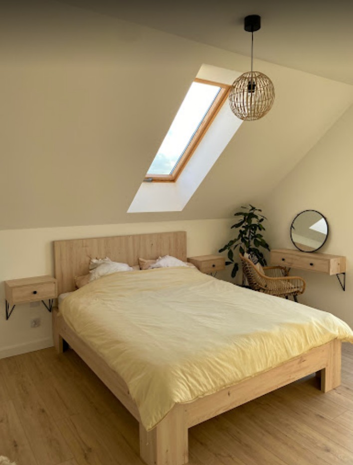 Jasna sypialnia na poddaszu z drewnianym łóżkiem, szafkami nocnymi na czarnych wspornikach i okrągłym lustrem nad toaletką, oświetlona lampą z abażurem z wikliny i oknem dachowym.