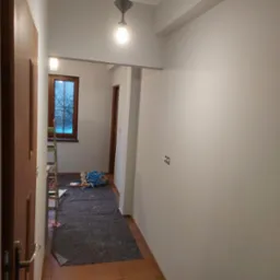 Korytarz w trakcie odświeżania: świeżo pomalowane ściany w odcieniach szarości, zabezpieczona podłoga, drabina oparta o okno z widokiem na drzewa bez liści za szybą.