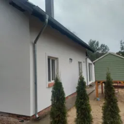 Boczny widok otynkowanego na biało domu z szarym dachem i rynnami, z widocznymi oknami w białych ramach, czarną lampą zewnętrzną, ceglaną podmurówką, trzema iglakami i zieloną szopą w tle.