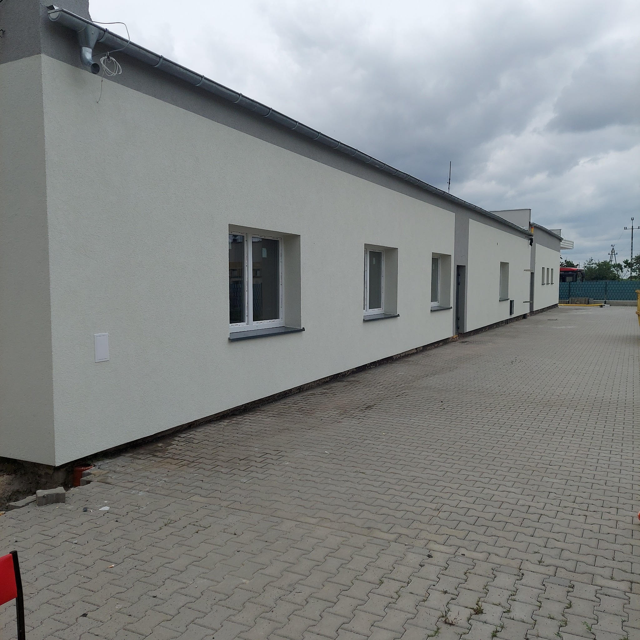 Świeżo wykończona elewacja budynku z widocznymi oknami i szarymi elementami wykończeniowymi, z chodnikiem z kostki brukowej na pierwszym planie i pochmurnym niebem w tle.