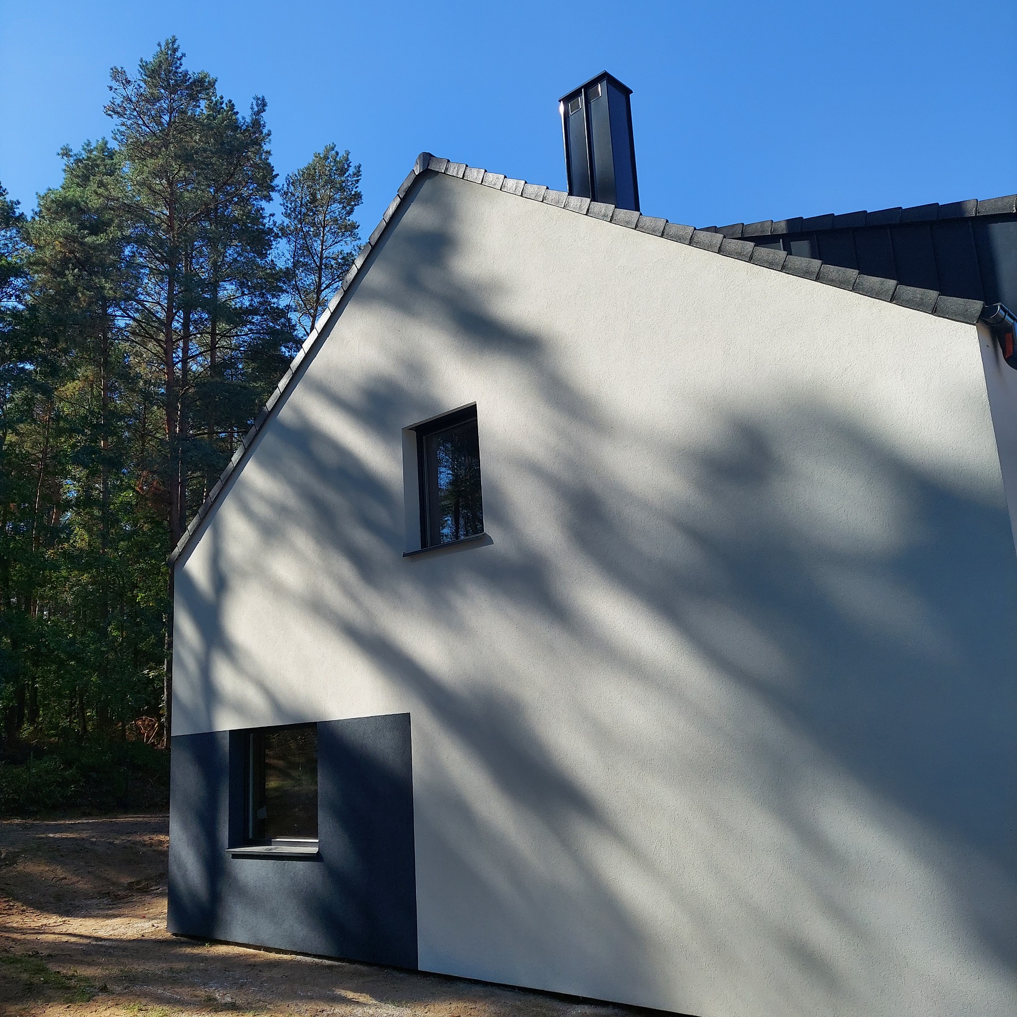 Nowoczesny dom jednorodzinny z jasnoszarą elewacją i ciemnoszarą podstawą, widoczne dwa okna i czarny komin na tle lasu i błękitnego nieba.