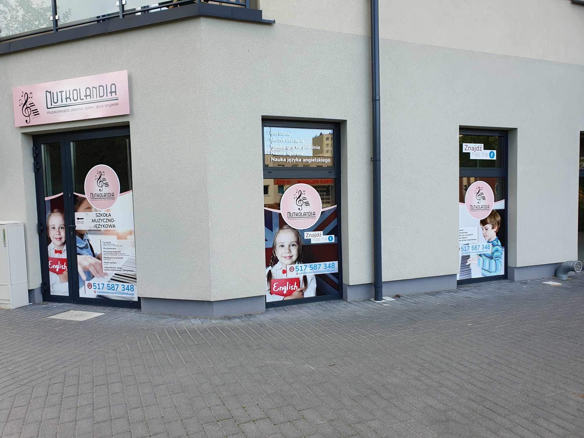 Fasada budynku szkoły muzyczno-językowej Nutkolandia z widocznymi plakatami reklamowymi w oknach i na drzwiach, prezentującymi ofertę zajęć muzycznych i języka angielskiego dla dzieci.