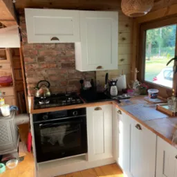 Drewniana kuchnia w rustykalnym stylu z białymi szafkami, ceglaną ścianą i piecem na drewno obok piekarnika.