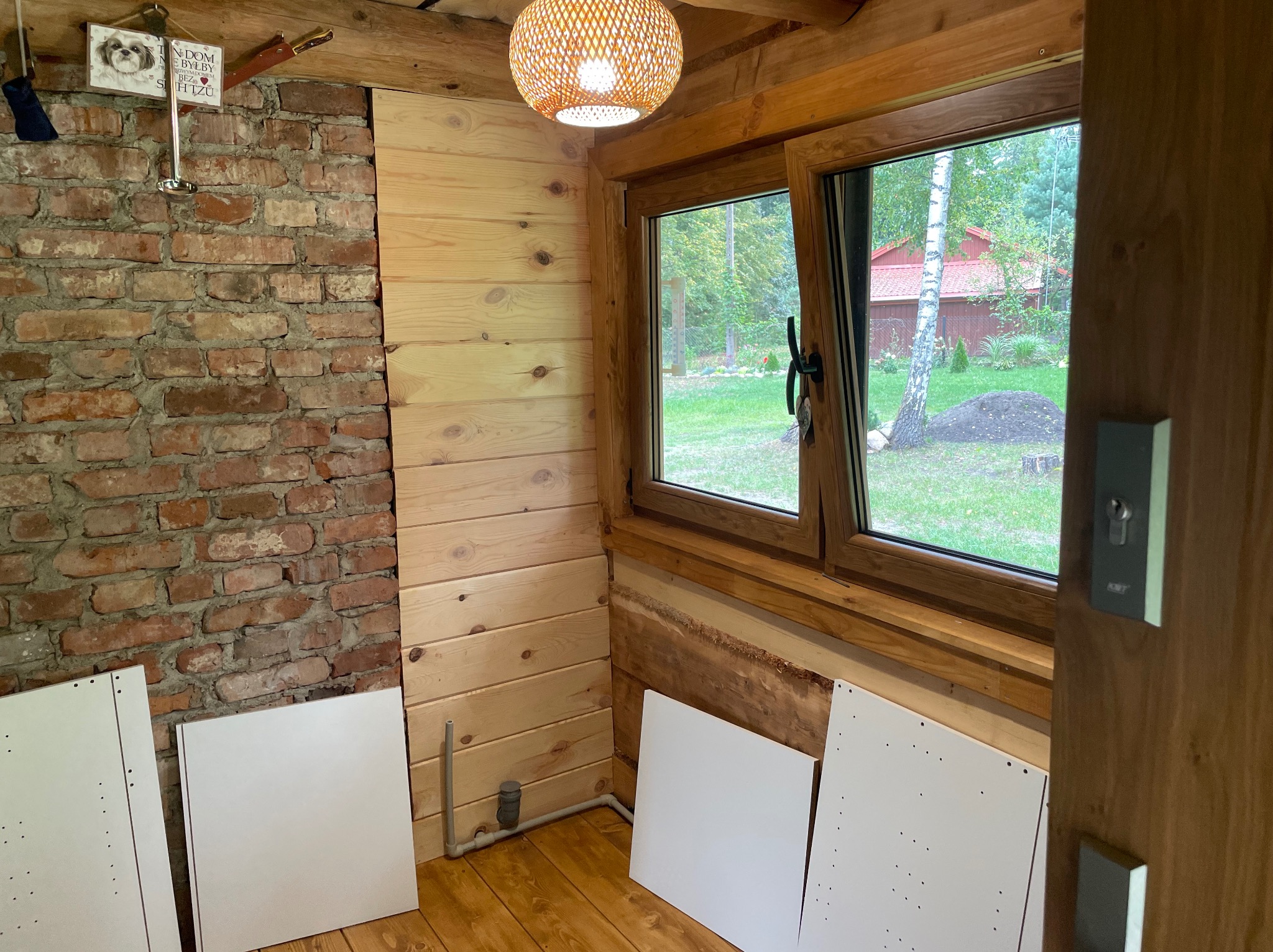 Wnętrze drewnianego domku z oknem na ogród, widoczne elementy mebli do montażu, ściana z cegły oraz drewniana boazeria
