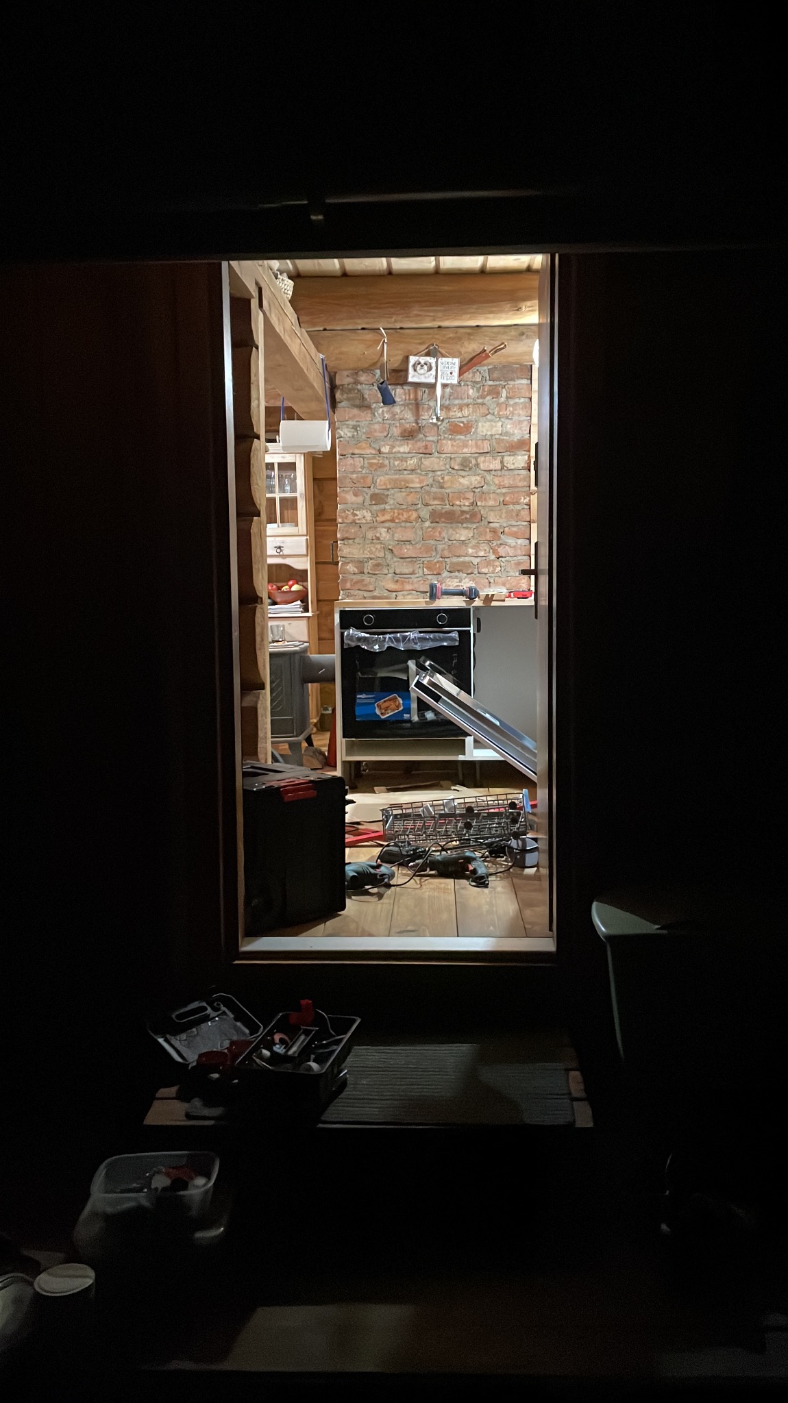 Wnętrze domu w trakcie remontu, widok przez drzwi: niedokończona zabudowa kuchenna z piekarnikiem, ceglana ściana, narzędzia na podłodze, słabe oświetlenie.