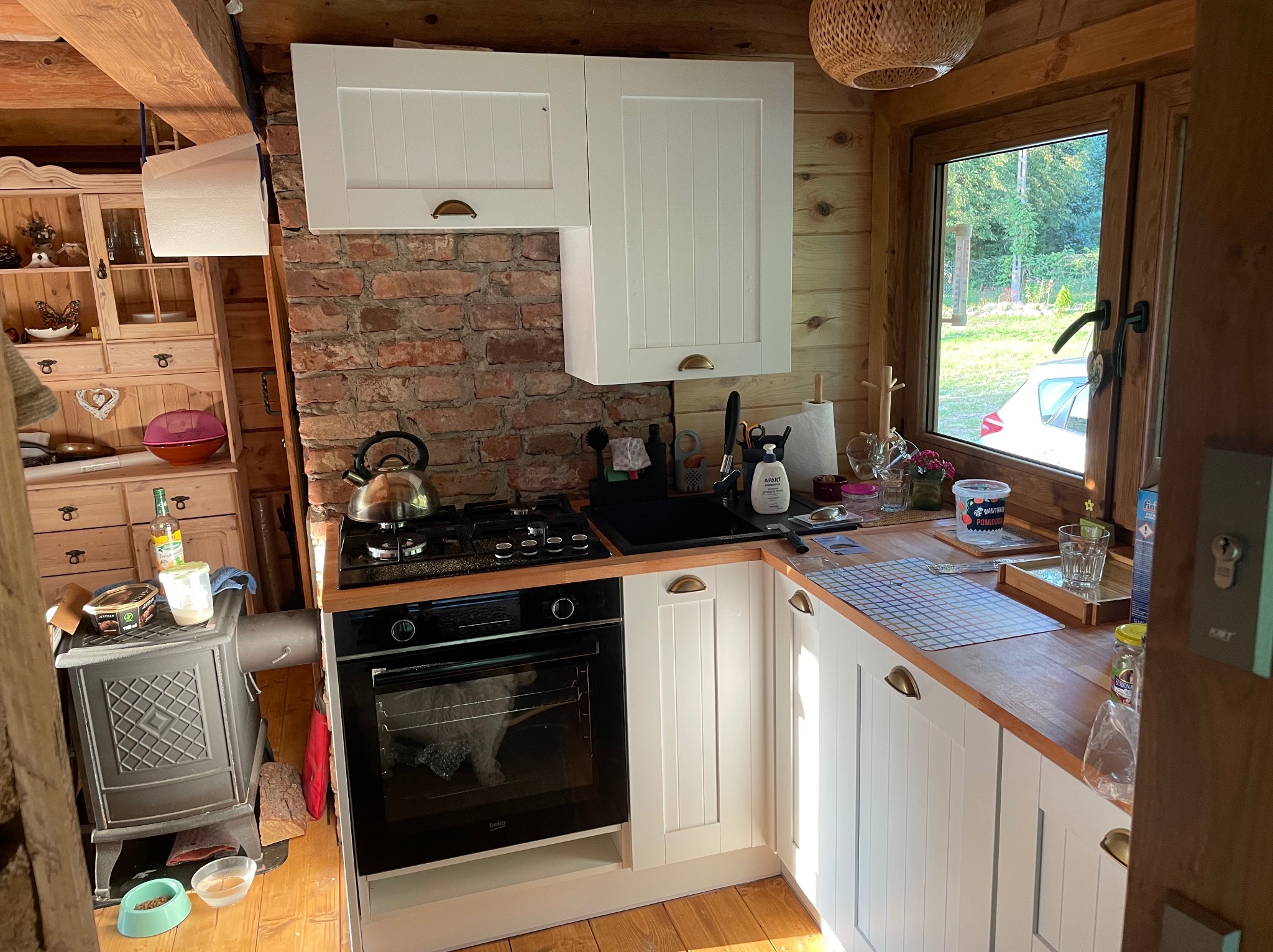 Drewniana kuchnia w rustykalnym stylu z białymi szafkami, ceglaną ścianą i piecem na drewno obok piekarnika.