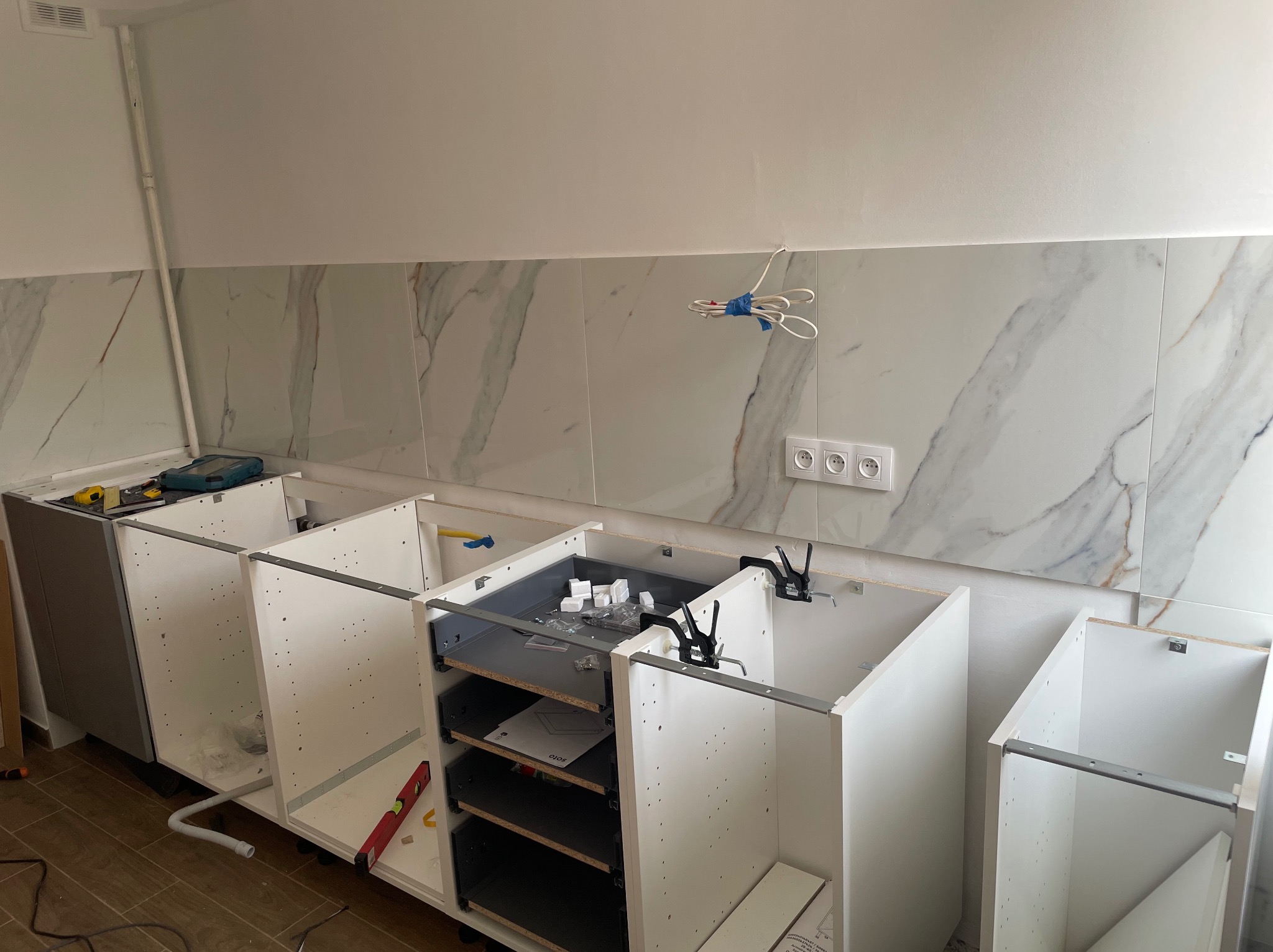 Fragment kuchni w trakcie montażu: białe szafki bez frontów, częściowo zamontowane szuflady, widoczne narzędzia i akcesoria montażowe na blacie, marmurowe płytki na ścianie, wystające przewody...