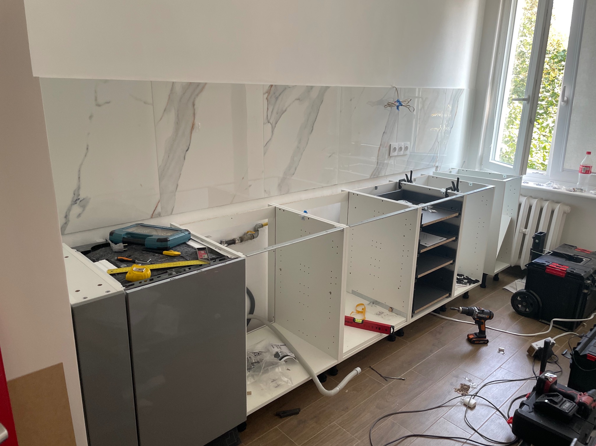 Wnętrze w trakcie instalacji białych szafek kuchennych z szarymi frontami, widoczne narzędzia, poziomica, wiertarka, skrzynki narzędziowe i płytki imitujące marmur na ścianie.