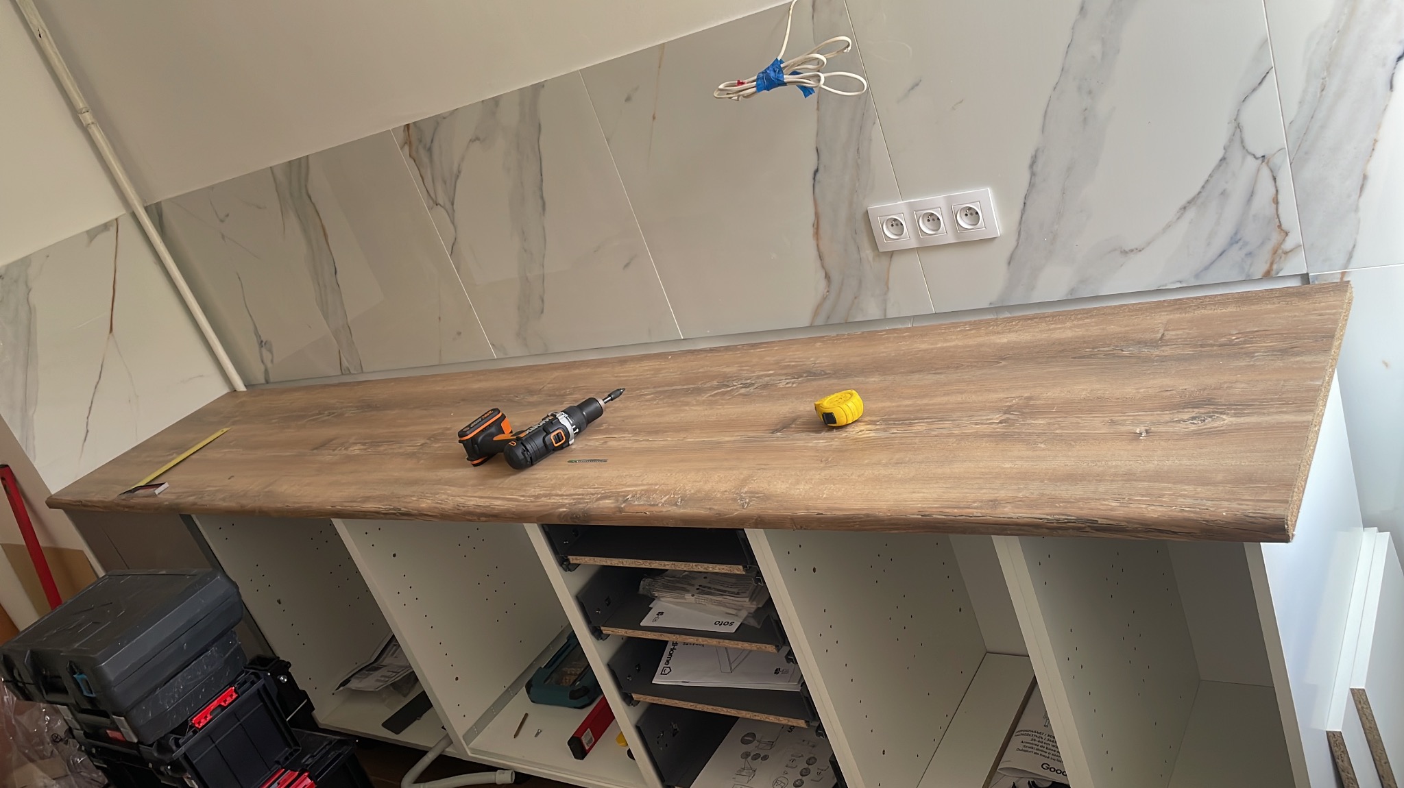 Blat drewnopodobny w trakcie montażu na białej szafce, widoczna wiertarko-wkrętarka akumulatorowa, miarka zwijana i poziomica, tło stanowi marmurowa ściana z gniazdkami.