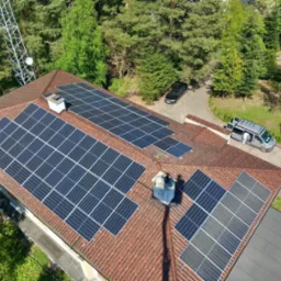 Widok z drona na dom z panelami fotowoltaicznymi na dachu z czerwonej dachówki, na podjeździe stoi srebrny samochód dostawczy z otwartymi drzwiami, w tle drzewa i wieża telekomunikacyjna.