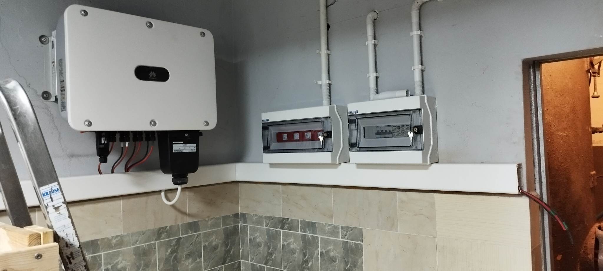 Zamontowany inwerter Huawei z podłączonymi przewodami, dwie skrzynki z bezpiecznikami na ścianie w pomieszczeniu z wykończeniem z płytek imitujących kamień, fragment drabiny z naklejką Krause.