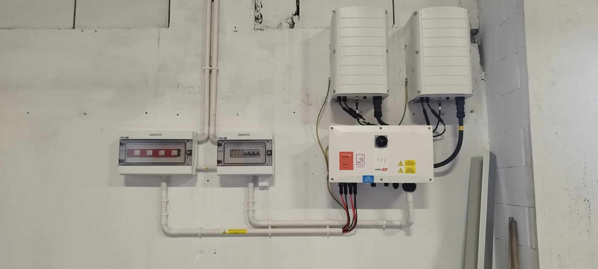 Dwa białe inwertery fotowoltaiczne zamontowane na ścianie, połączone kablami z białą skrzynką z ostrzegawczymi naklejkami, poniżej widoczne dwie skrzynki elektryczne z bezpiecznikami i okablowaniem...