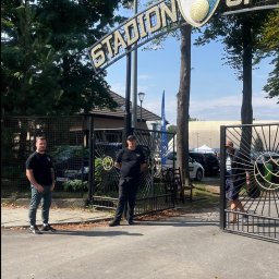 Grupa Najemnik Sp z o.o - Dwóch ochroniarzy w czarnych uniformach stoi przy wejściu na Stadion KS Borek, z widocznym logo klubu i fragmentem trybun w tle.