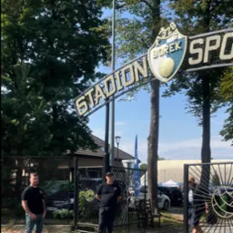 Dwóch ochroniarzy w czarnych uniformach stoi przy otwartej bramie wejściowej na Stadion Sportowy KS Borek z herbem klubu i napisem 'Stadion'.