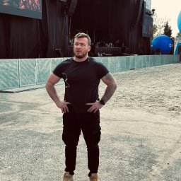 Grupa Najemnik Sp z o.o - Mężczyzna w czarnym ubraniu z tatuażem na ramieniu stoi na tle sceny koncertowej z ekranami i głośnikami, prawdopodobnie pracownik ochrony.