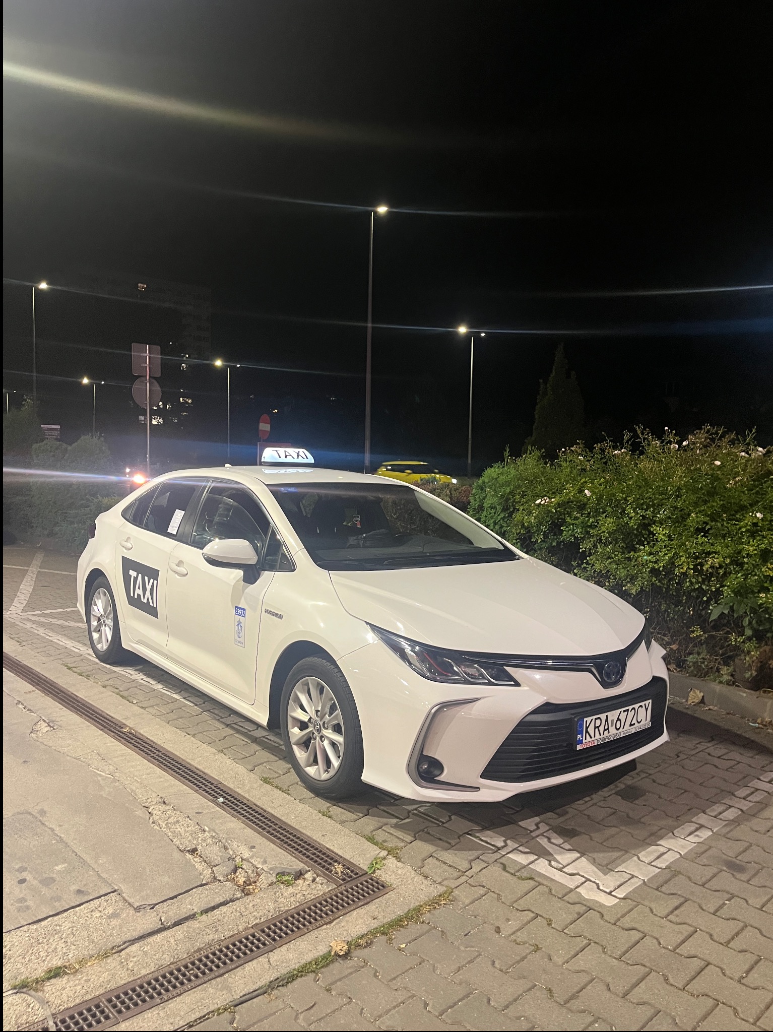 Biała Toyota Corolla Taxi zaparkowana nocą na oświetlonym parkingu, widoczny znak taxi na dachu i logo na drzwiach. Tablica rejestracyjna KRA-672CY.