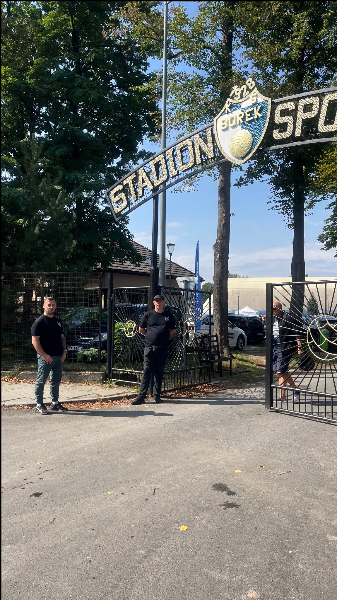 Dwóch ochroniarzy w czarnych uniformach stoi przy wejściu na Stadion KS Borek, z widocznym logo klubu i fragmentem trybun w tle.