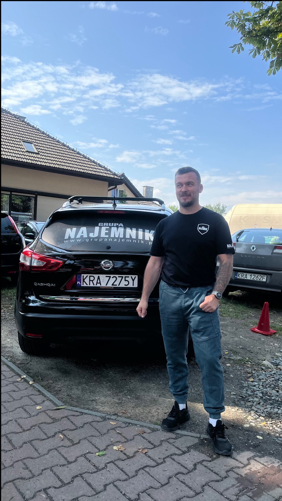 Mężczyzna stoi obok czarnego Nissana Qashqai z naklejką 'Grupa Najemnik' na tylnej szybie. Widoczny budynek w tle i fragment zaparkowanego auta.