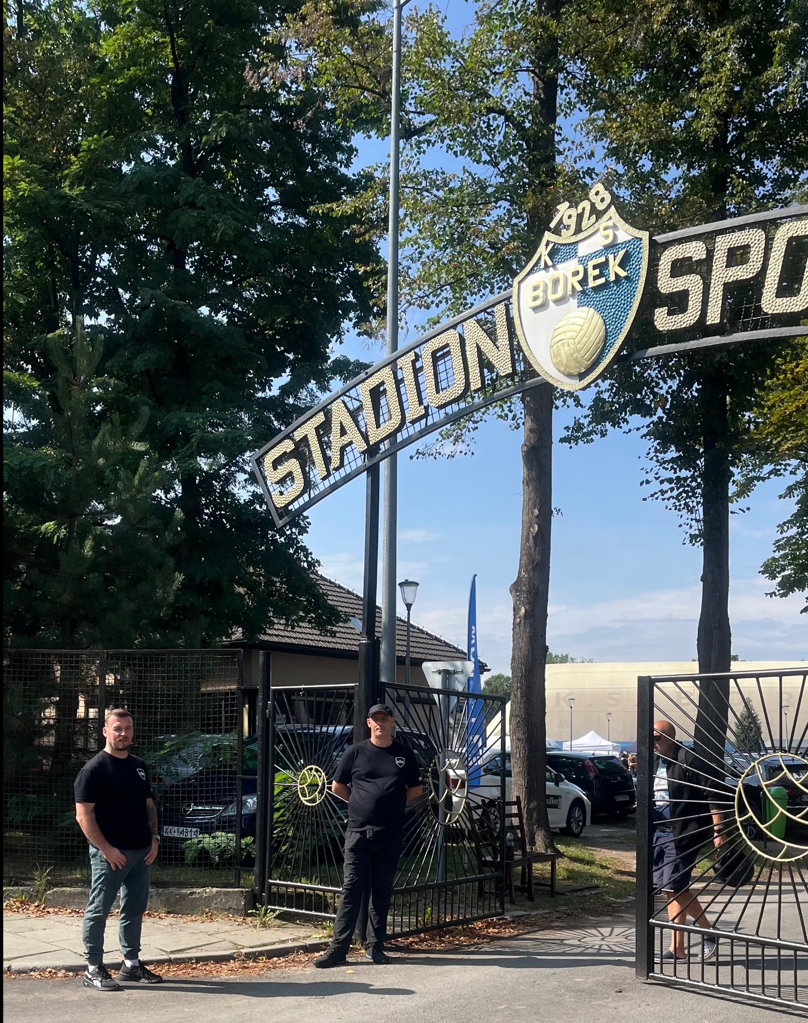 Dwóch ochroniarzy w czarnych uniformach stoi przy otwartej bramie wejściowej na Stadion Sportowy KS Borek z herbem klubu i napisem 'Stadion'.