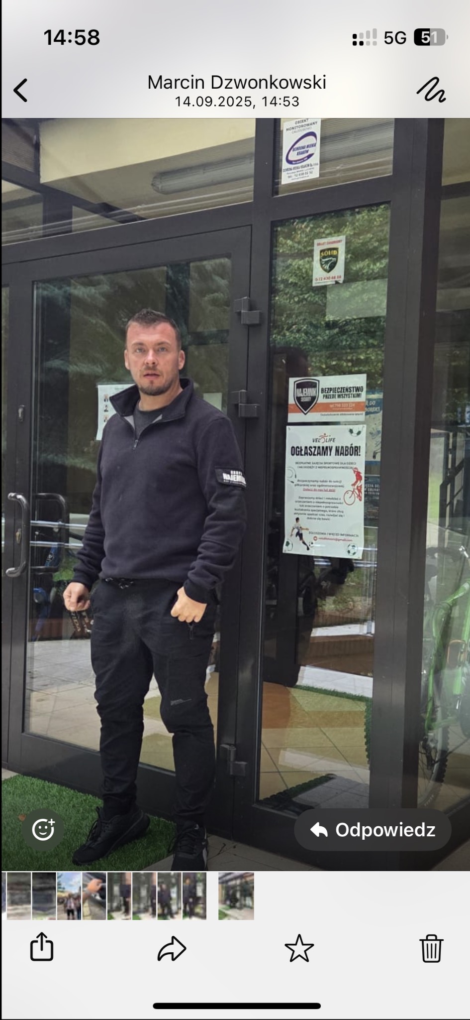 Mężczyzna w czarnym stroju firmy ochroniarskiej stoi przed wejściem do budynku z naklejkami agencji ochrony. Widoczne logo firmy ochroniarskiej na odzieży.