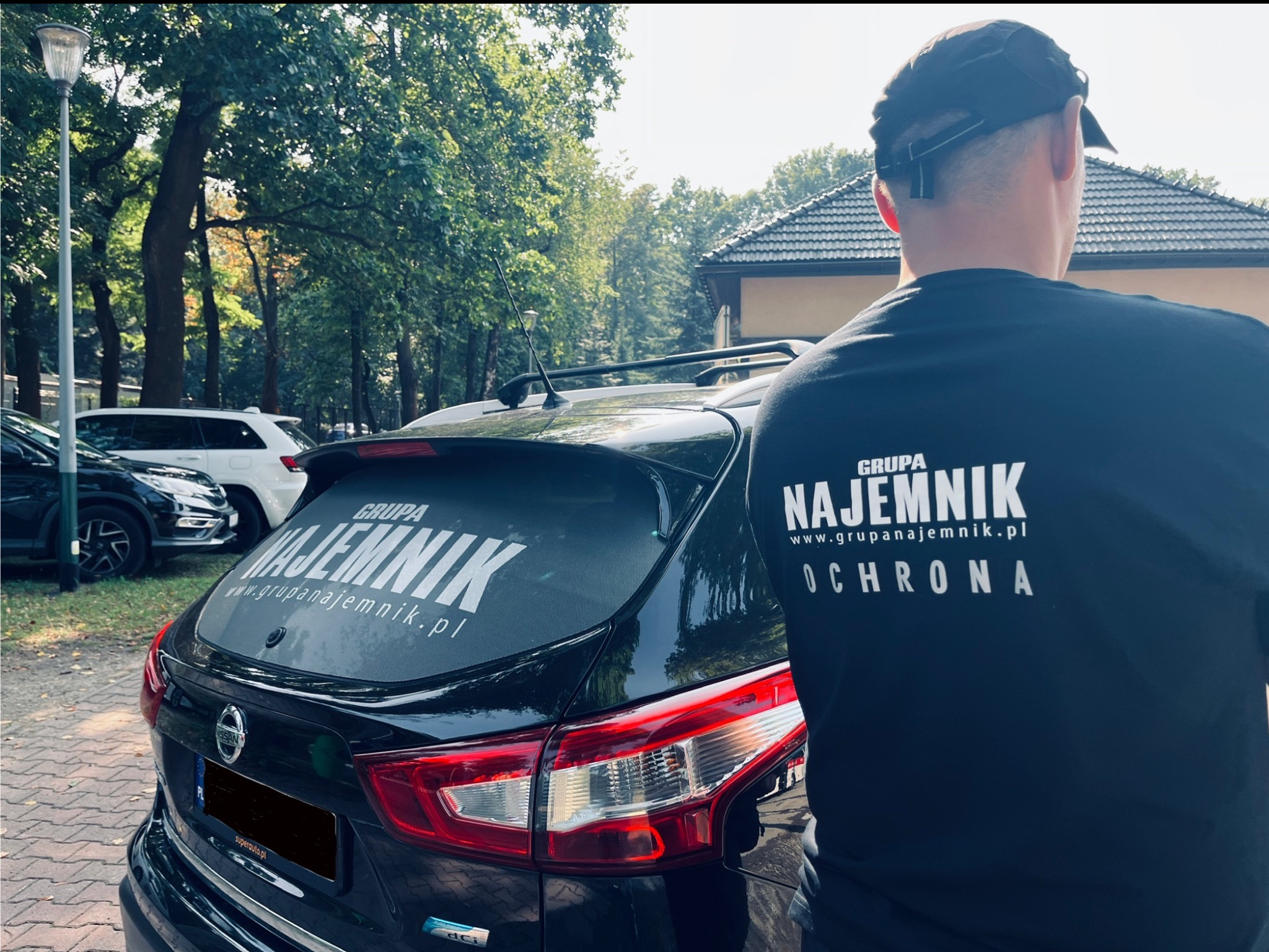 Czarny Nissan Qashqai z naklejką 'Grupa Najemnik Ochrona' na tylnej szybie, obok stoi mężczyzna w koszulce z tym samym napisem, na tle drzew i budynku.