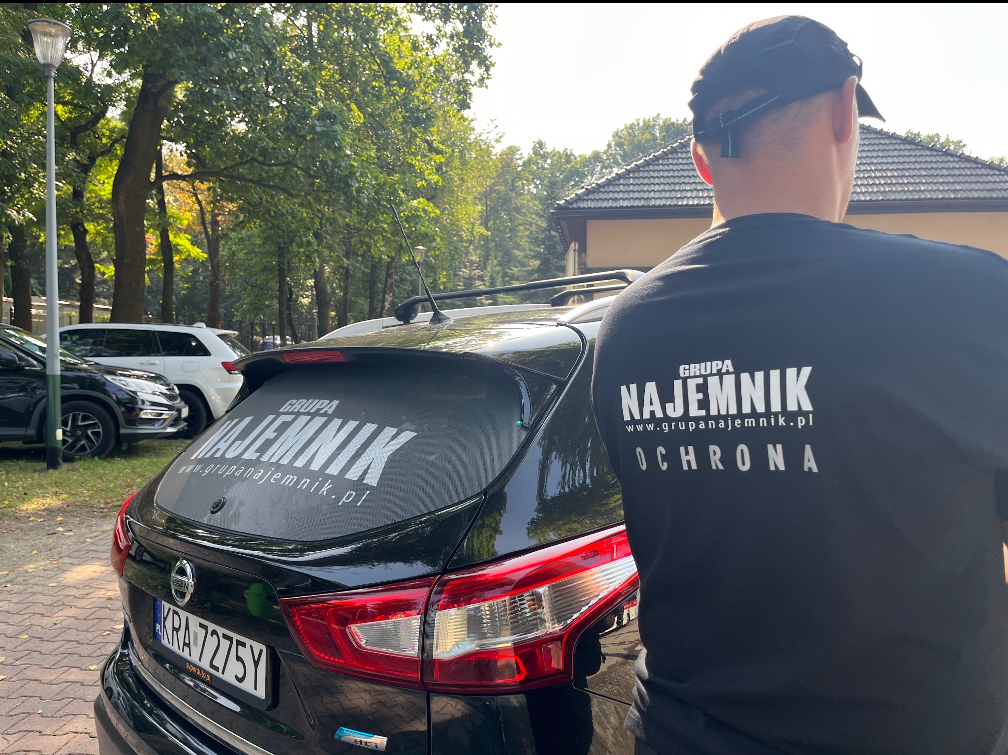 Czarny samochód z naklejką 'Grupa Najemnik Ochrona' na tylnej szybie, obok stoi mężczyzna w koszulce z tym samym napisem, na tle zieleni.