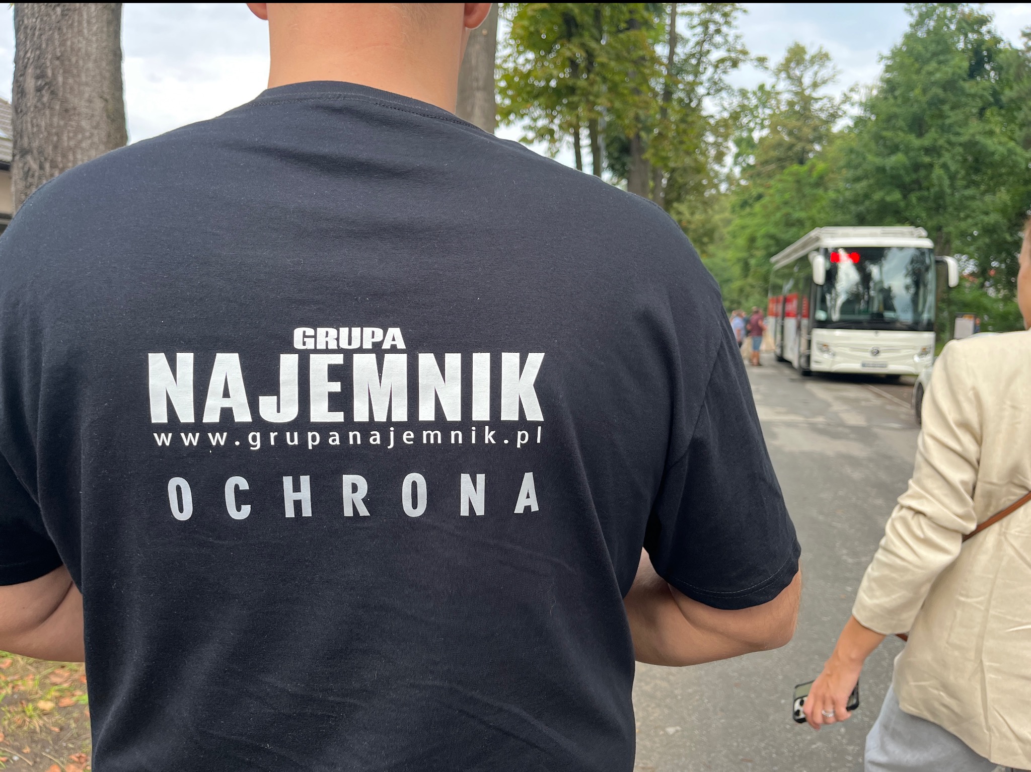 Czarna koszulka z napisem 'GRUPA NAJEMNIK www.grupanajemnik.pl OCHRONA' na plecach mężczyzny. W tle autobus i drzewa, sugerujące plenerową lokalizację.