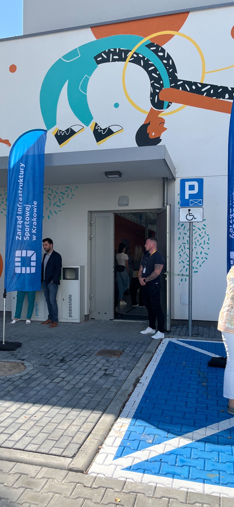 Wejście do budynku z muralem sportowym, flaga z logo i napisem 'Zarząd Infrastruktury Sportowej w Krakowie', ochroniarz w czarnym ubraniu i biale sneakersy, oraz osoby wchodzące do budynku.