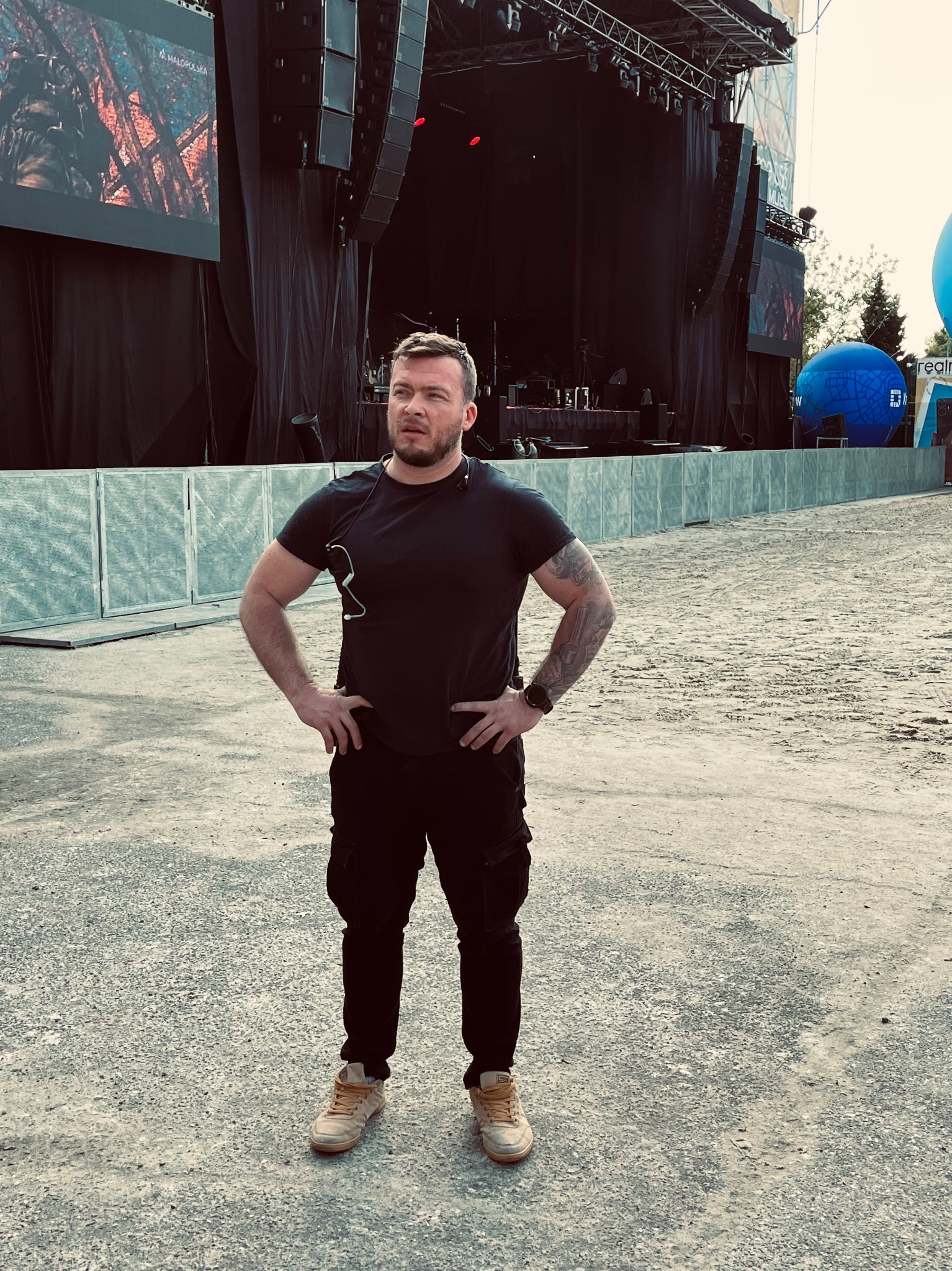 Mężczyzna w czarnym ubraniu z tatuażem na ramieniu stoi na tle sceny koncertowej z ekranami i głośnikami, prawdopodobnie pracownik ochrony.
