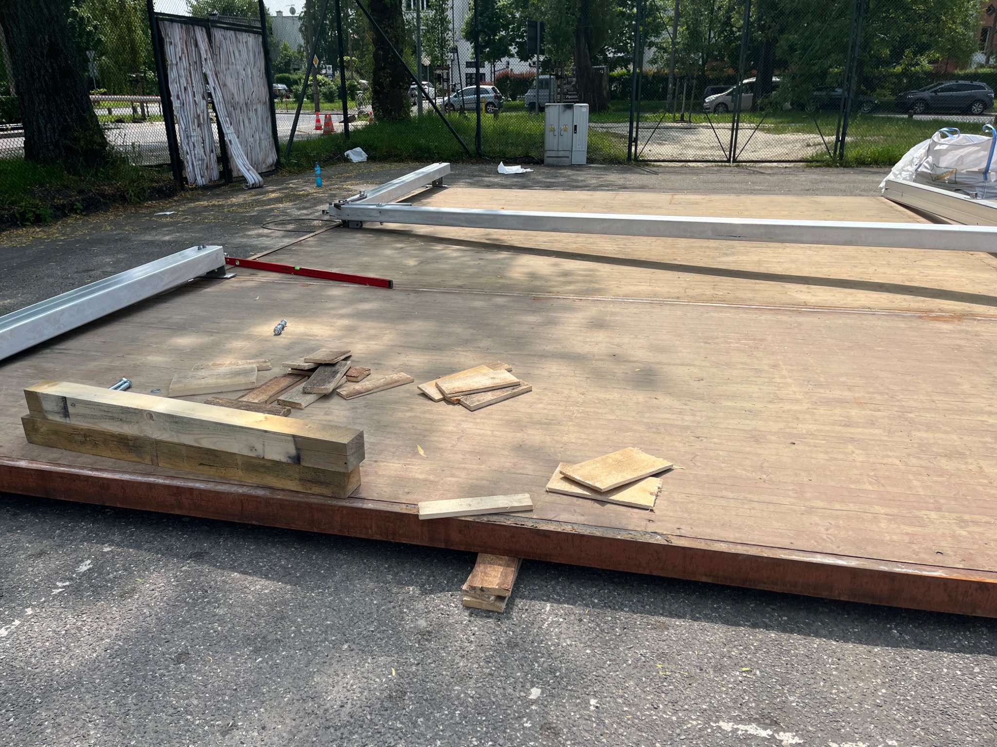 Podest z metalową ramą w trakcie budowy na zewnątrz, z poziomnicą i fragmentami drewna, otoczony zielenią i elementami infrastruktury miejskiej.