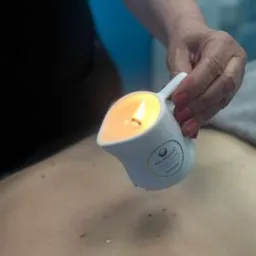 Dłoń terapeuty wylewa ciepły wosk ze specjalnego dzbanka na plecy klienta podczas zabiegu relaksacyjnego. Widoczne drobinki wosku na skórze.